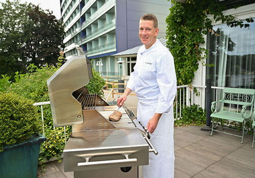 Koch grillt Fleisch auf einer Terrasse vor einem Gebäude.