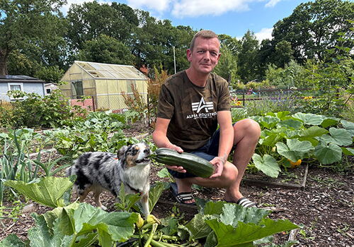 Mann mit Zucchini und Hund im Garten vor Gewächshaus