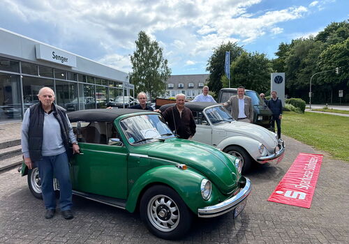 Gruppe posiert mit klassischen VW-Käfern vor Autohaus.