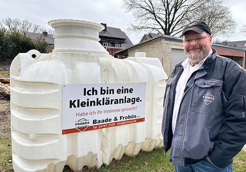 Mann steht neben einer weißen Kleinkläranlage mit Schild.