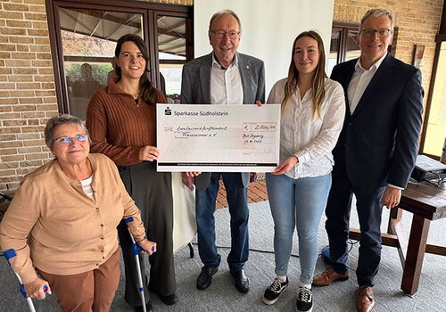 Fünf Personen halten einen großen Scheck in einem Raum.