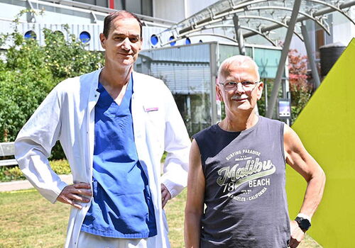 Zwei Männer stehen draußen; einer trägt einen Arztkittel.