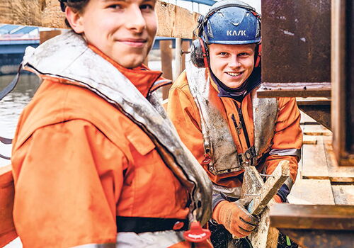 Zwei Arbeiter in Schutzkleidung auf einer Baustelle am Wasser.
