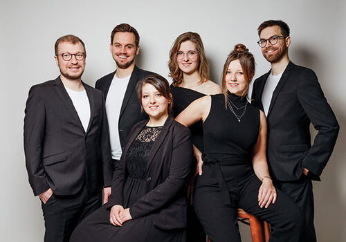 Sechs Personen in eleganter Kleidung posieren zusammen.