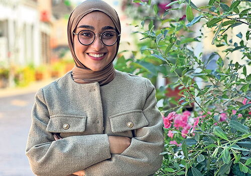 Frau mit Kopftuch und Brille lächelt vor Blumen.