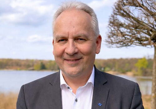 Mann im Anzug lächelt im Freien vor einem See.