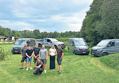 Gruppe posiert auf Wiese vor drei grauen Fahrzeugen.