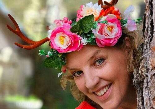 Frau mit Blumen- und Geweihkrone lächelt hinter einem Baum hervor.