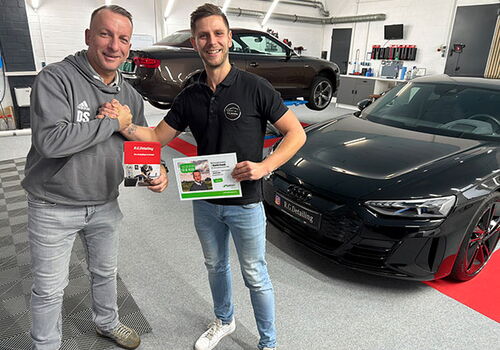 Zwei Männer neben einem Auto, halten Zertifikate.