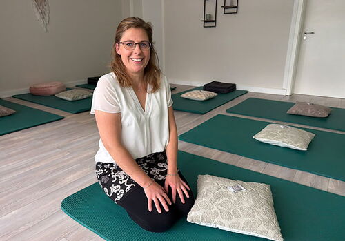 Frau sitzt lächelnd auf Matte in hellem Yogaraum.