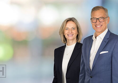 Zwei Personen in Businesskleidung lächeln vor unscharfem Hintergrund.