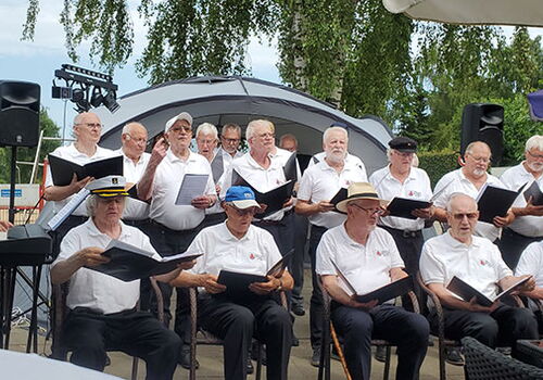 Männerchor singt im Freien, Bäume im Hintergrund.