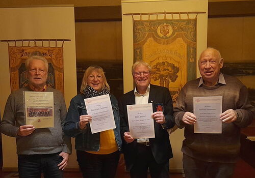 Vier Personen halten Urkunden vor gemusterten Bannern.