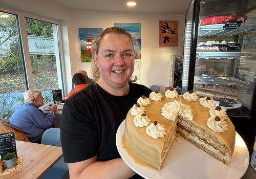 Frau hält Torte im Café, im Hintergrund Gäste und Kunst.