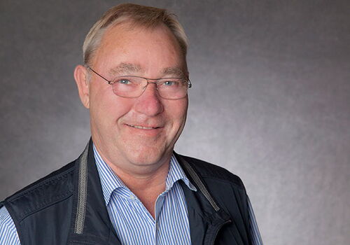 Mann mit Brille und Weste lächelt vor grauem Hintergrund.