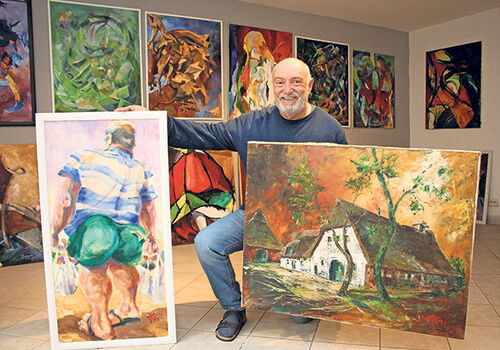 Mann zeigt zwei Gemälde, sitzt vor einer Kunstwand.