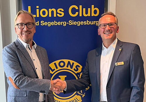 Zwei Männer schütteln sich vor einem Lions Club Banner die Hand.