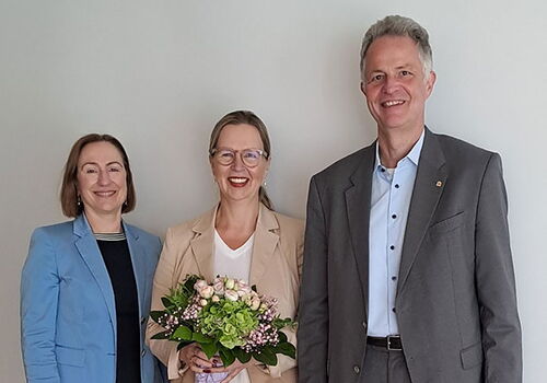 Drei Personen stehen lächelnd nebeneinander mit Blumenstrauß.