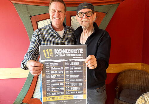 Zwei Männer halten ein Konzertplakat in einem Raum.