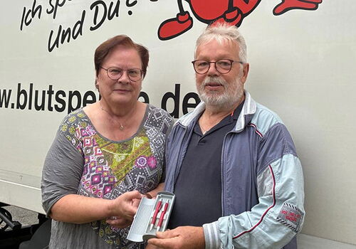 Zwei Personen vor einem Blutspende-Lkw mit Geschenk.