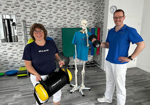 Zwei Personen mit Hanteln vor einem Skelett in einem Fitnessraum.
