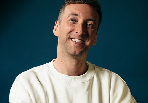 Lächelnde Person mit verschränkten Armen vor blauem Hintergrund.