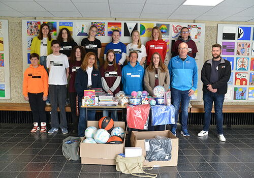 Schulgruppe mit Sportgeräten und Geschenken in einer Aula.