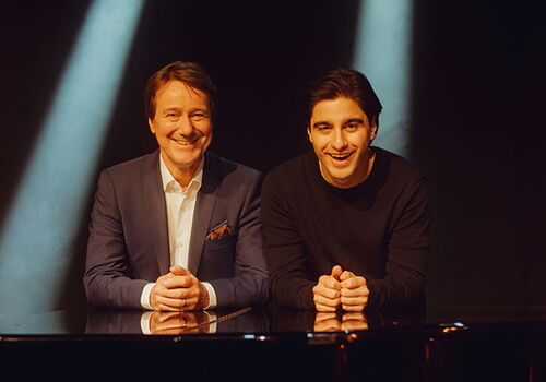 Zwei Männer lächeln im Scheinwerferlicht hinter einem Klavier.