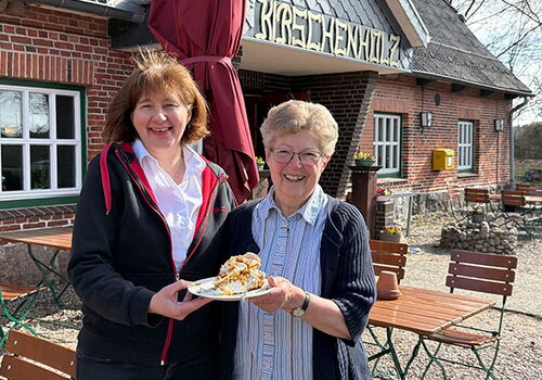 Zwei Frauen halten Kuchen vor einem Backsteinhaus.