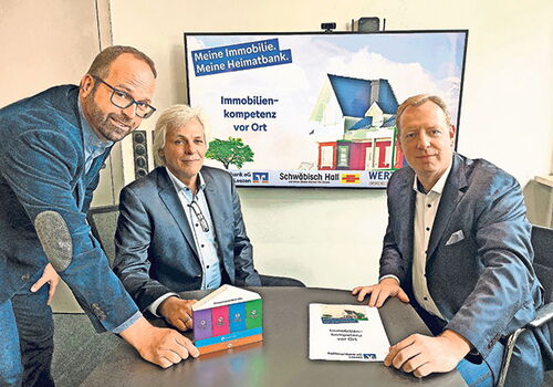 Drei Männer an einem Tisch vor Immobilien-Werbung.
