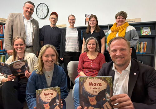Gruppe posiert mit dem Buch 'Mara flattert los' in einer Bibliothek.