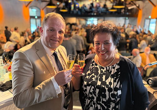 Zwei Personen stoßen mit Sektgläsern an, im Hintergrund feiernde Menge.