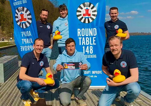 Gruppe von Männern mit Enten an einem See vor Bannern.