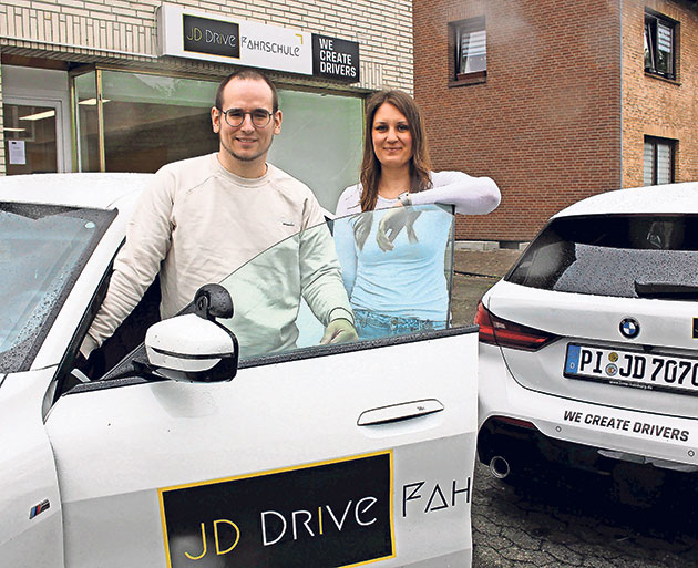 Fahrschule JD DRIVE in der Stadt Zwei Personen vor einer Fahrschule mit Autos im Hintergrund.