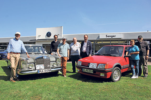 Gruppe posiert mit Oldtimern vor Autohaus.