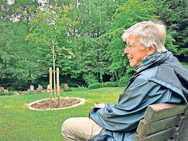 Person sitzt auf einer Bank vor einem Baum auf einem Friedhof.