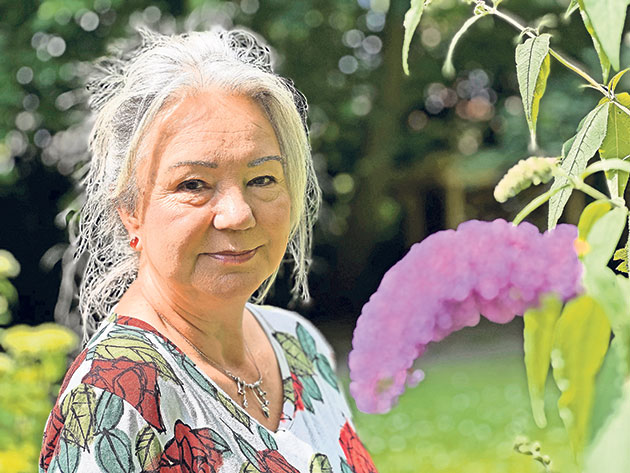 Ältere Frau im Garten mit lila Blüte im Vordergrund.