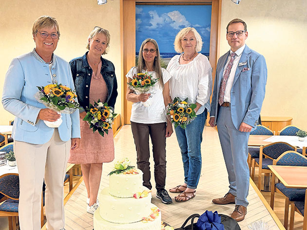 Gruppe mit Blumensträußen bei Feier mit großer Torte. Fünf Personen posieren mit Blumensträußen vor einer großen Torte.