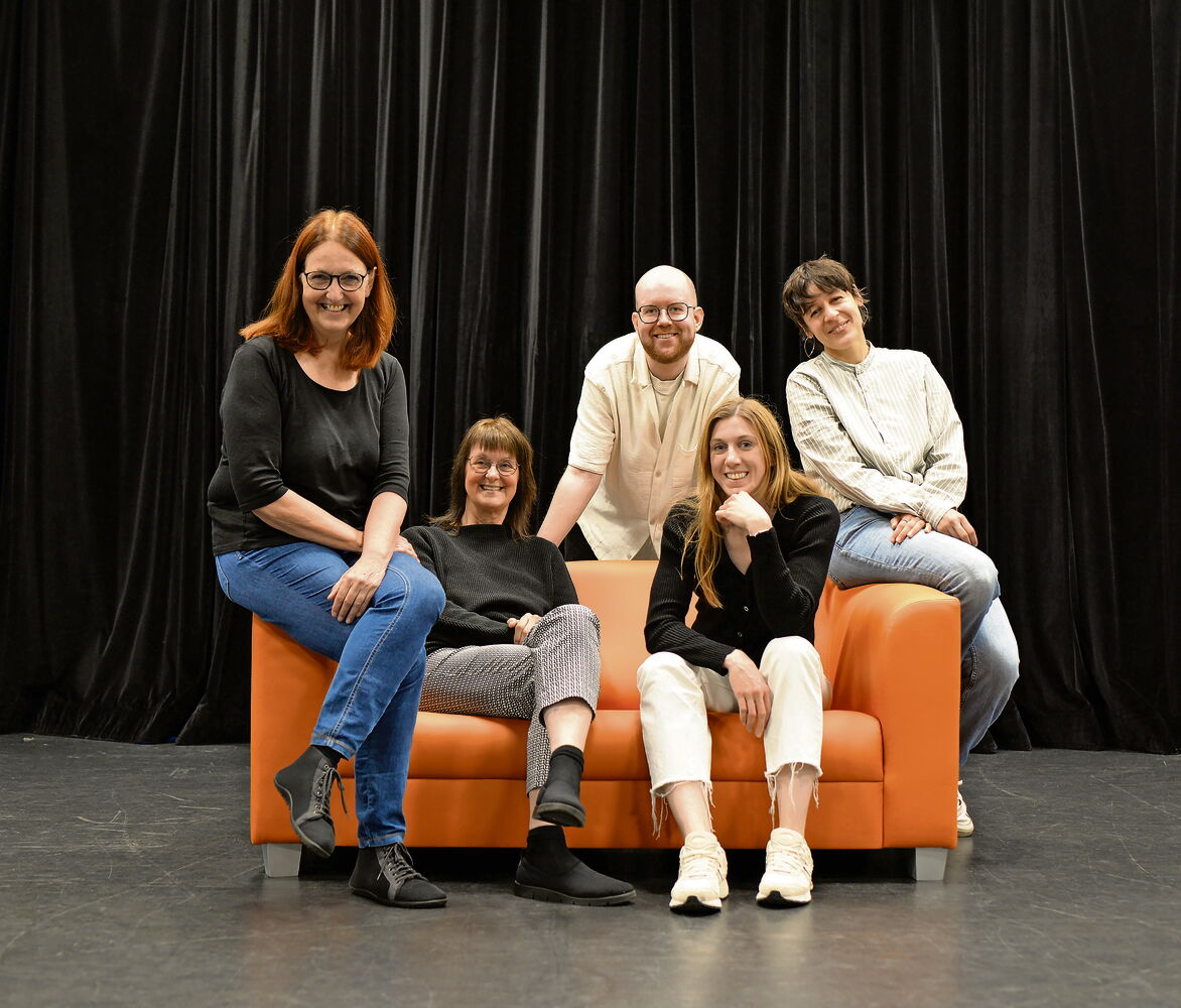 Gruppe auf Sofa vor schwarzem Vorhang Fünf Personen sitzen auf einem orangefarbenen Sofa.