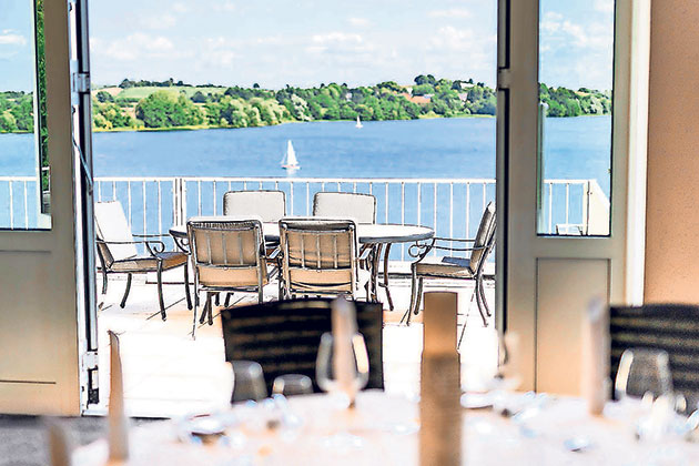 Terrasse mit Seeblick und Segelboot im Hintergrund. Blick von Terrasse auf See mit Segelboot und Bäumen.