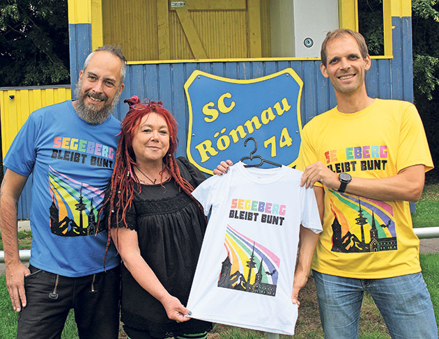 Drei Personen halten ein buntes T-Shirt vor einem SC Rönnau Schild.