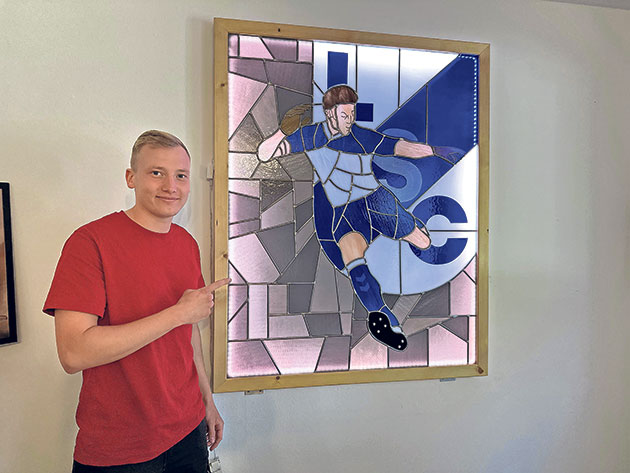 Person neben einem Fußballmosaik in Blau Mann zeigt auf ein Mosaik mit Fußballspieler in blauem Trikot.