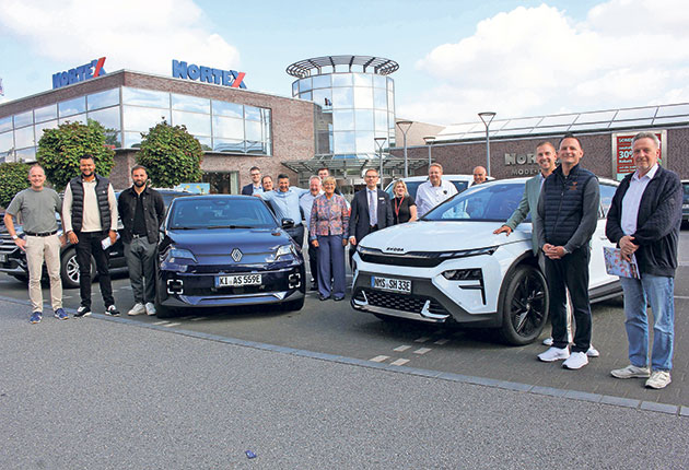 Menschen und Autos vor einem Geschäft Gruppe posiert neben zwei Autos vor Gebäude.