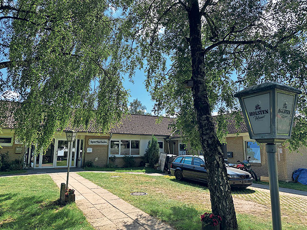 Sportlerheim im Grünen Sportlerheim mit Bäumen und parkendem Auto bei Sonnenschein.