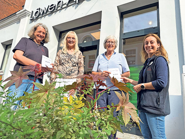 Vier Personen vor einer Apotheke mit Pflanzen im Vordergrund.
