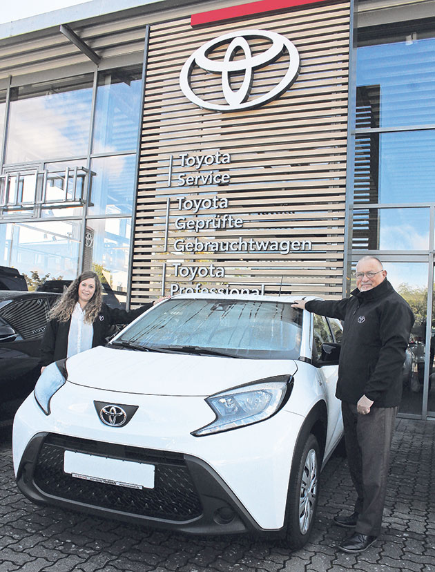 Zwei Personen stehen neben einem weißen Toyota vor einem Autohaus.