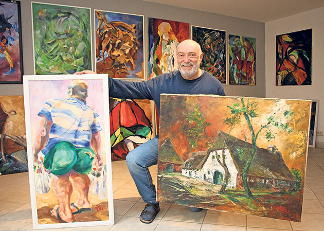 Künstler mit zwei Gemälden im Atelier Mann zeigt zwei Gemälde, sitzt vor einer Kunstwand.