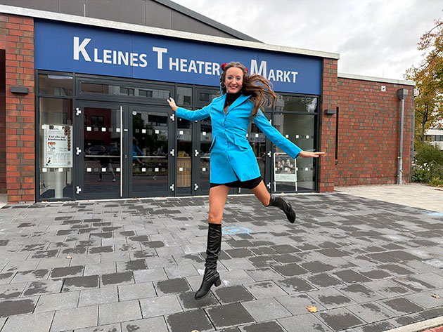 Frau in blauem Mantel vor Theatergebäude Frau springt vor dem Kleinen Theater Markt.