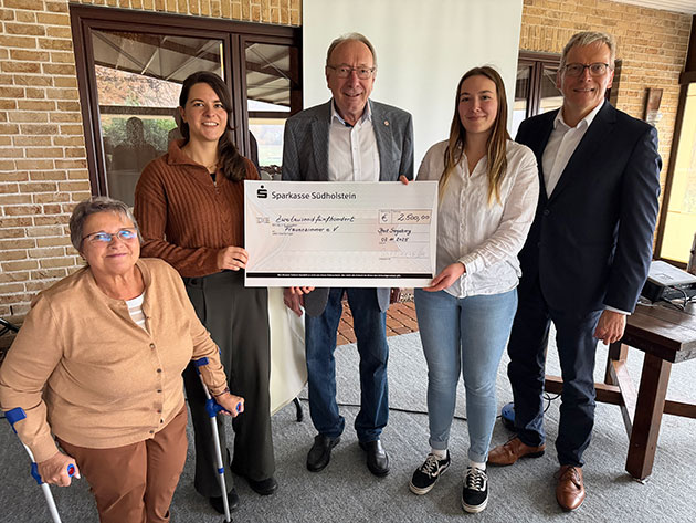 Fünf Personen halten einen großen Scheck in einem Raum.