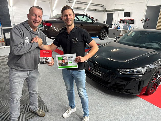 Zwei Männer neben einem Auto, halten Zertifikate.